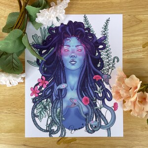 Pode incluir: Uma ilustração digital de Medusa, uma figura mitológica grega, com cobras como cabelo. Seu rosto é azul com reflexos rosados e está cercada por folhagem verde e flores rosas. A assinatura da artista, "Emma", está no canto inferior direito.