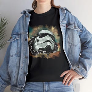 Pode incluir: Camiseta preta com um design de capacete de Stormtrooper entrelaçado com uma cobra. O gráfico é definido contra um fundo abstrato colorido. A camisa é usada com uma jaqueta jeans azul.