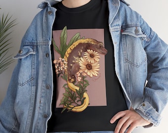 Könnte beinhalten: Schwarzes T-Shirt mit einer detaillierten Illustration eines Geckos mit gelben und braunen Markierungen, umgeben von Sonnenblumen, Blättern und einer Banane. Das Design befindet sich auf einem mauvefarbenen Hintergrund. Das Shirt wird mit einer blauen Jeansjacke getragen.