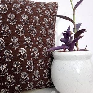 K&ouml;nnte beinhalten: Ein braunes Zierkissen mit wei&szlig;em Blumenmuster liegt neben einem wei&szlig;en, gesprenkelten Keramiktopf mit einer lila Pflanze. Das Kissen hat eine gesteppte Textur und ein sich wiederholendes Blumendesign.