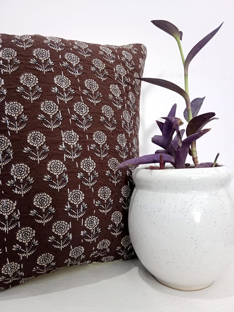 K&ouml;nnte beinhalten: Ein braunes Zierkissen mit wei&szlig;em Blumenmuster liegt neben einem wei&szlig;en, gesprenkelten Keramiktopf mit einer lila Pflanze. Das Kissen hat eine gesteppte Textur und ein sich wiederholendes Blumendesign.