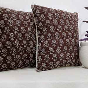 K&ouml;nnte beinhalten: Zwei quadratische Zierkissen mit einem dunkelbraunen Blumenmuster. Die Kissen haben einen wei&szlig;en Rand und liegen auf einer wei&szlig;en Oberfl&auml;che. Ein kleiner wei&szlig;er Topf mit einer Pflanze steht neben den Kissen.