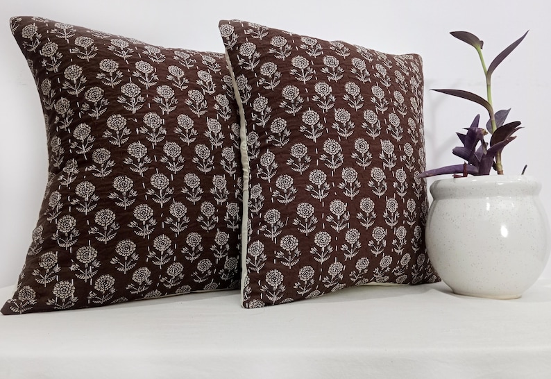K&ouml;nnte beinhalten: Zwei quadratische Zierkissen mit einem dunkelbraunen Blumenmuster. Die Kissen haben einen wei&szlig;en Rand und liegen auf einer wei&szlig;en Oberfl&auml;che. Ein kleiner wei&szlig;er Topf mit einer Pflanze steht neben den Kissen.