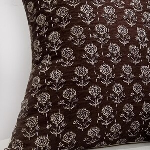 K&ouml;nnte beinhalten: Ein braunes Zierkissen mit einem sich wiederholenden wei&szlig;en Blumenmuster. Das quadratische Kissen besteht aus einem strukturierten Stoff. Das florale Design umfasst stilisierte Blumen und Bl&auml;tter.