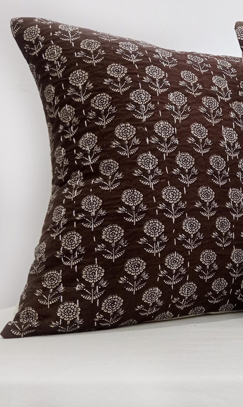 K&ouml;nnte beinhalten: Ein braunes Zierkissen mit einem sich wiederholenden wei&szlig;en Blumenmuster. Das quadratische Kissen besteht aus einem strukturierten Stoff. Das florale Design umfasst stilisierte Blumen und Bl&auml;tter.