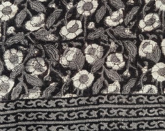 May include: A close-up of a textile with a floral pattern. The design features white flowers and gray leaves on a dark brown background. The bottom edge has a decorative border and tassels.