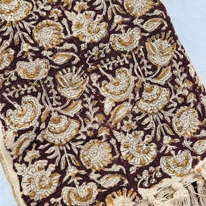 May include: A folded textile with a repeating floral pattern in shades of burgundy, cream, and gold. The edges are finished with a fringe. The design features stylized flowers and leaves, creating a decorative and textured appearance.
