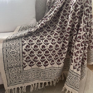 May include: A cream-coloured throw blanket with a dark brown floral pattern and black border. The blanket has fringe on all sides and is draped over a sofa.