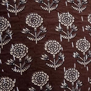 K&ouml;nnte beinhalten: Dunkelbrauner Stoff mit einem sich wiederholenden Muster aus wei&szlig;en Blumenmotiven. Jede Blume hat mehrere Bl&uuml;tenbl&auml;tter und ist mit einem Stiel mit Bl&auml;ttern verbunden. Der Stoff hat ein gestepptes Aussehen mit vertikalen Stichlinien.