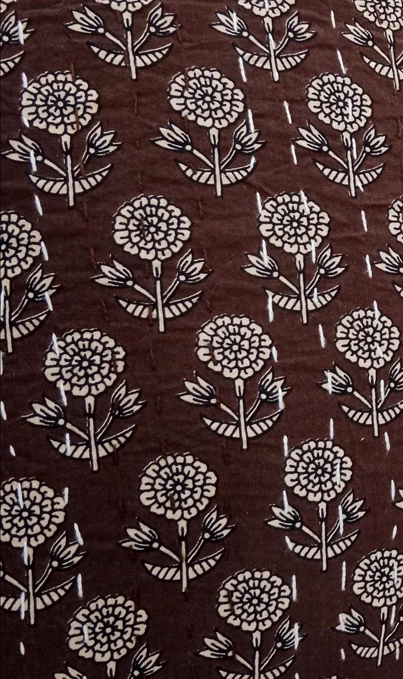 K&ouml;nnte beinhalten: Dunkelbrauner Stoff mit einem sich wiederholenden Muster aus wei&szlig;en Blumenmotiven. Jede Blume hat mehrere Bl&uuml;tenbl&auml;tter und ist mit einem Stiel mit Bl&auml;ttern verbunden. Der Stoff hat ein gestepptes Aussehen mit vertikalen Stichlinien.