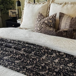 May include: A bed featuring a light-colored, fine-striped duvet cover and two large ruffled pillows. The bedding includes a dark brown throw blanket with a cream-colored floral pattern, a square floral accent pillow, and a rectangular textured pillow.