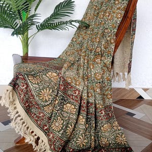 May include: A floral patterned throw blanket draped over a wooden chair. The blanket features a repeating design of flowers in shades of green, brown, and red. The edges are trimmed with a cream-coloured fringe. A potted plant is visible in the background.