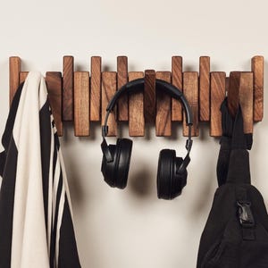May include: A wooden wall-mounted coat rack with multiple pegs of varying heights. A black and white striped jacket, black headphones, and a black sling bag hang from the rack. The rack is made of dark brown wood.