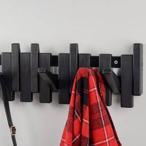 May include: Black wooden wall-mounted coat rack with multiple hooks. A red and black plaid shirt and a black leather strap hang from the hooks.