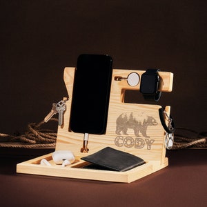 May include: A wooden desk organizer with multiple compartments. It features a phone holder, watch stand, and key hooks. The organizer has a bear and forest engraving with the name "CODY". A black wallet, keys, and a smartwatch are visible.