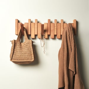 Peut inclure: Un porte-manteau mural en bois avec un pull marron et un sac tiss&eacute; accroch&eacute;s. Un collier en or avec un pendentif en forme d'&eacute;toile est &eacute;galement accroch&eacute; au porte-manteau.