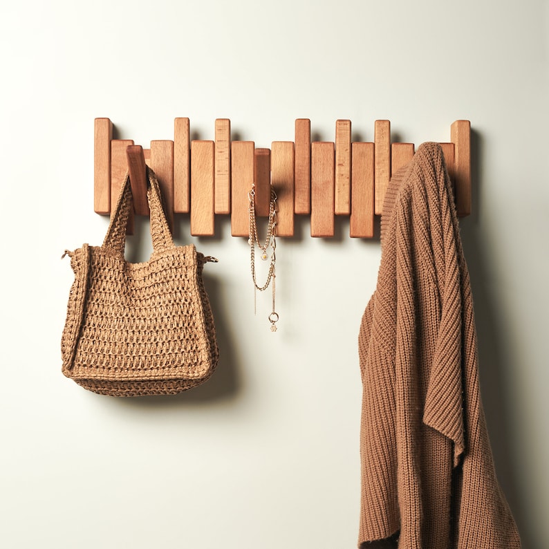 Peut inclure: Un porte-manteau mural en bois avec un pull marron et un sac tiss&eacute; accroch&eacute;s. Un collier en or avec un pendentif en forme d'&eacute;toile est &eacute;galement accroch&eacute; au porte-manteau.