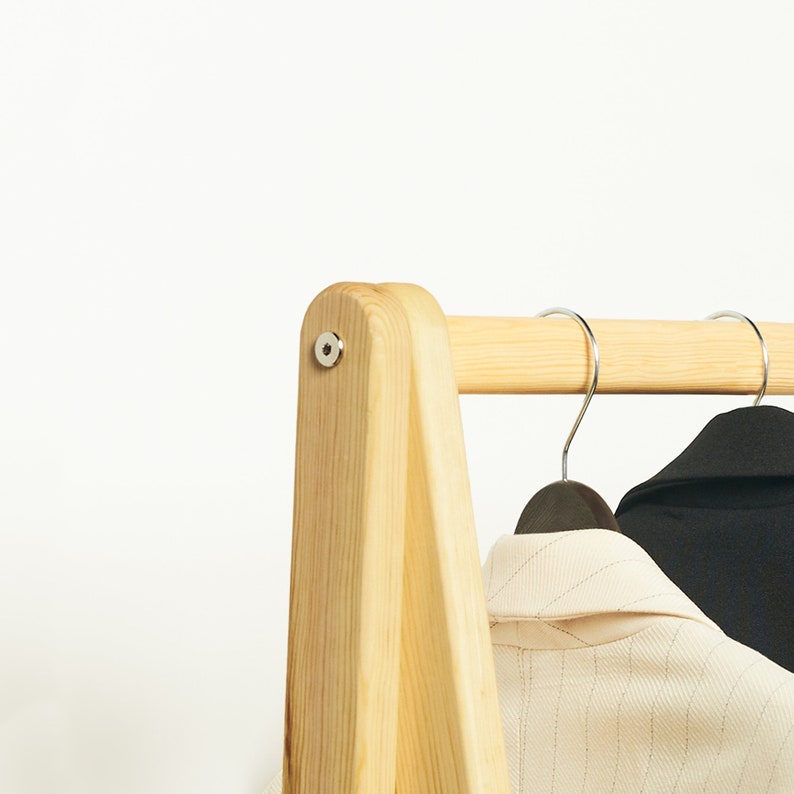 May include: A wooden clothing rack with a natural finish. The rack has a triangular base and a horizontal bar for hanging clothes. Two hangers with clothes are hanging on the bar.