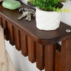 May include: A dark brown wooden wall-mounted shelf featuring a flat top surface and a series of vertical wooden slats underneath. A small white geometric pot with a green plant, a set of metal keys, and a light green earbud case sit on the shelf.