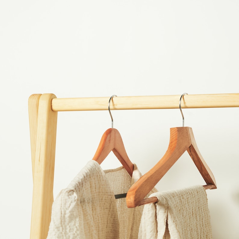 May include: Two wooden hangers with a natural finish hanging on a wooden rack. The hangers are made of a light brown wood and have a simple design. A white garment is hanging on the hangers.