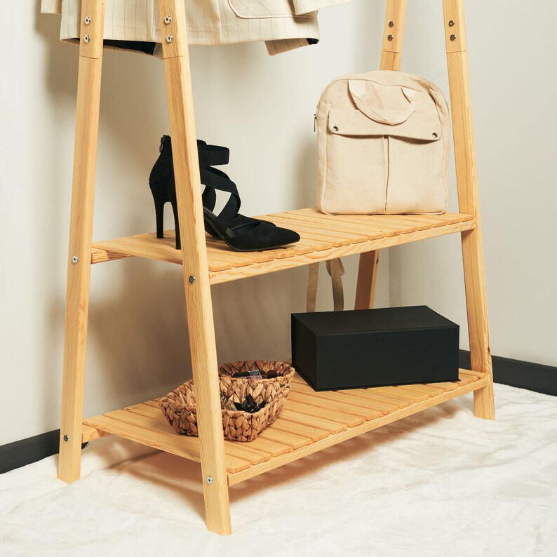 May include: A wooden clothing rack with two shelves. The top shelf has a beige backpack and a pair of black heeled shoes. The bottom shelf has a black box and two woven baskets.