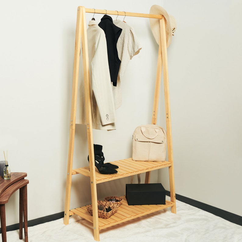 May include: A wooden clothing rack with two shelves. The rack is made of natural wood and has a simple, modern design. It is holding a white blazer, a black sweater, a white shirt, a hat, a bag, and shoes.