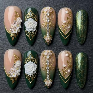 May include: A set of ten artificial nails with a variety of designs. The nails feature a combination of nude, green, and gold colors. Some nails have 3D floral accents, while others have ornate gold filigree and rhinestone details. One nail is a glittery green.