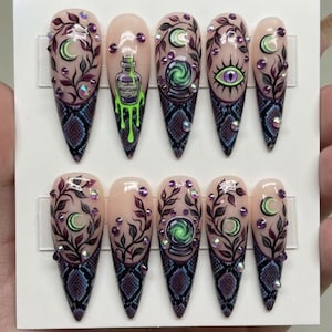 May include: A set of ten stiletto-shaped press-on nails featuring a nude base with a blue and purple snakeskin pattern at the tips. Designs include hand-painted vines, crescent moons, a potion bottle labeled Nightshade, crystal balls, and an eye.