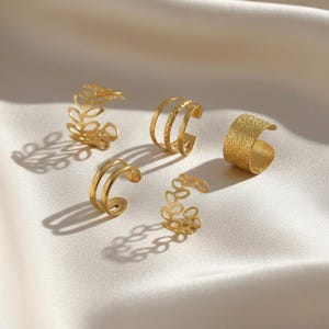 Puede incluir: Cinco anillos dorados de varios diseños se muestran sobre una superficie blanca. Los anillos incluyen una banda texturizada, bandas dobles y diseños en forma de hoja. Los anillos están hechos de un material metálico brillante.