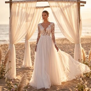 May include: A white wedding dress with a V-neckline and long sleeves, featuring delicate floral lace detailing. The dress has a flowing tulle skirt and is worn on a beach with a wooden archway and sheer white curtains. The overall aesthetic is romantic and ethereal.