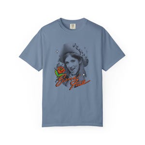 May include: A slate blue short-sleeve t-shirt featuring a black and white portrait of a person wearing a hat. Below the portrait is a colorful red and green rose illustration and the orange cursive text Sierra Ferrell with small star accents.