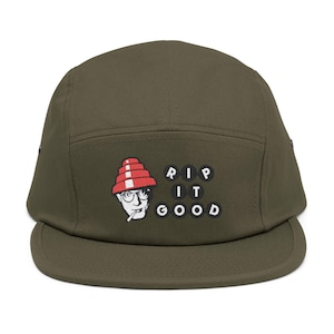 May include: Olive green five-panel cap with a flat brim. The cap features an embroidered design of a cartoon character wearing a red and white hat, glasses, and a cigarette. The text "RIP IT GOOD" is embroidered on the cap.