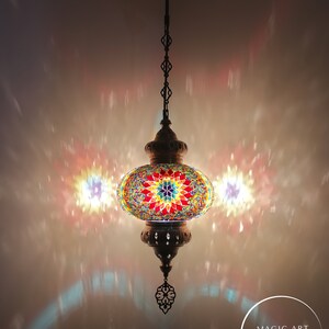May include: A colorful mosaic Turkish lamp with a starburst design hangs from a chain. The lamp is lit and casts a warm glow on the surrounding area. The text "Magic Art Turkish Lamp Mosaic Lights" is visible in the lower right corner of the image.