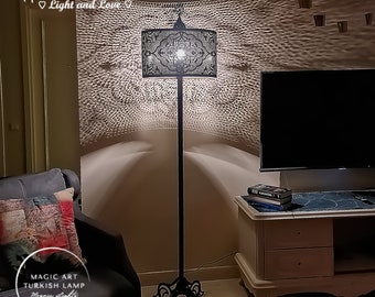 Op de afbeelding: Een zwarte staande lamp met een gepatenteerde lampenkap werpt ingewikkelde schaduwen op de muur. De lampenkap heeft een delicaat, ingewikkeld ontwerp en wordt van binnenuit verlicht. De lamp staat op een zwarte voet met sierlijke details. De lamp bevindt zich in een woonkamer met een bank en een tv-meubel op de achtergrond.