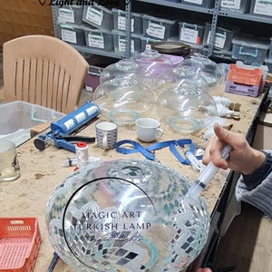 May include: A close-up of a glass mosaic Turkish lamp being assembled. The lamp is round and has a silver and turquoise mosaic design. The text "MAGIC ART TURKISH LAMP" is visible on the lamp.