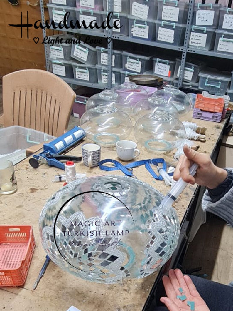 May include: A close-up of a glass mosaic Turkish lamp being assembled. The lamp is round and has a silver and turquoise mosaic design. The text "MAGIC ART TURKISH LAMP" is visible on the lamp.