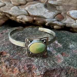 May include: A silver cuff bracelet with a textured band and a square centerpiece. The centerpiece features a light green oval stone set within a square frame with decorative accents. The bracelet is displayed on a textured, multi-colored surface.