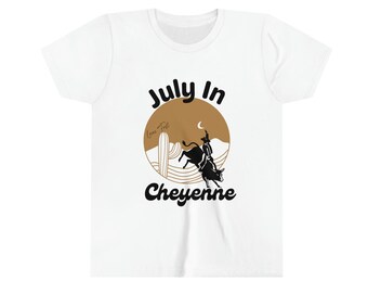 Puede incluir: Camiseta blanca con un diseño de rodeo. El diseño incluye un vaquero montando un toro, una luna creciente y las palabras "July In Cheyenne" en negro y marrón. También incluye un cactus y un paisaje desértico.