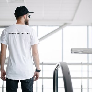 Puede incluir: Camiseta blanca con el texto "SORRY IF YOU CAN'T SEE" en negro. La camiseta la lleva una persona con gorra y gafas de sol negras. La persona est&aacute; de espaldas a la c&aacute;mara.