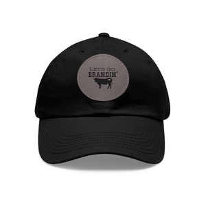 Puede incluir: Gorra de béisbol negra con visera curva. Un parche circular en la parte delantera muestra el texto "Lets Go Brandin'" sobre una silueta de toro. El parche es gris, y el texto y el toro son negros.