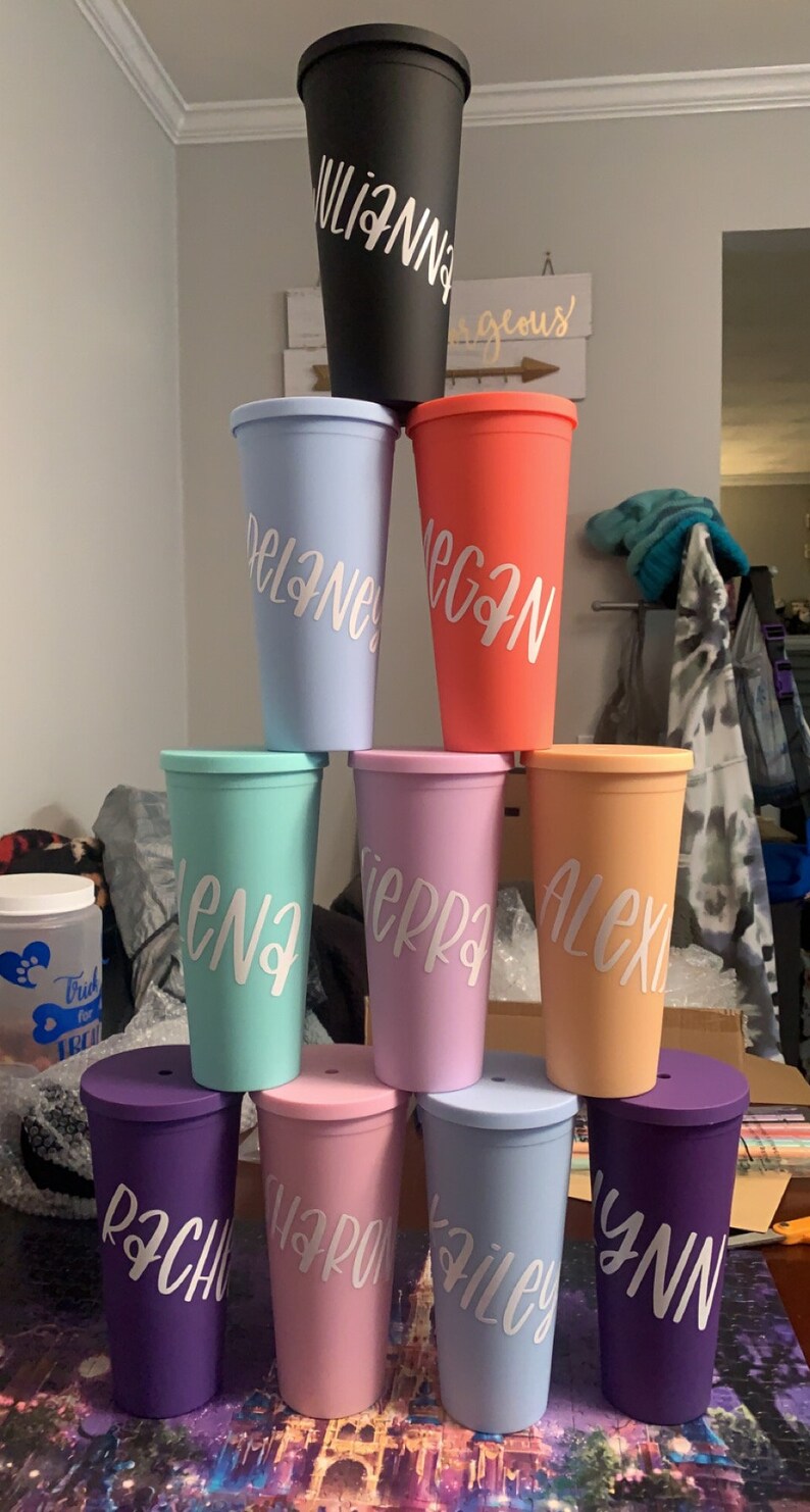 May include: A stack of nine colourful reusable tumblers with names written on them in white lettering. The tumblers are arranged in a pyramid shape, with the largest tumbler at the top and the smallest tumblers at the bottom.