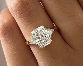 Puede incluir: Un anillo de oro con un gran diamante de talla cojín en el centro y dos diamantes más pequeños de forma pera a cada lado.
