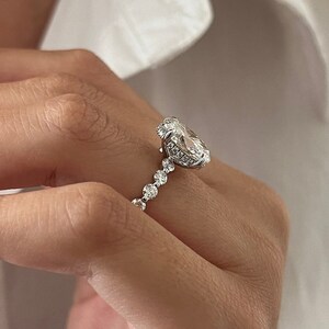K&ouml;nnte beinhalten: Ein silberner Ring mit einem gro&szlig;en, oval geschliffenen Diamanten als Mittelstein und kleineren runden Diamanten entlang des Rings. Der Ring ist vor einem wei&szlig;en Hintergrund abgebildet.