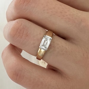 Może przedstawiać: Złoty pierścionek z diamentem o szlifie szmaragdowym.