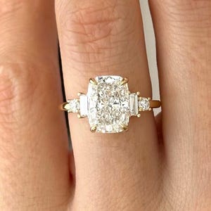 May include: A gold ring featuring a large, rectangular-cushion cut diamond center stone, flanked by smaller baguette diamonds. The ring is set on a delicate gold band, showcasing a classic and elegant design. The ring is shown on a hand with red nail polish.