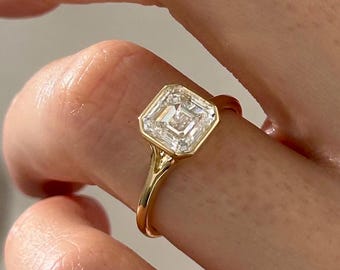 May include: A gold ring featuring a square-cut, clear gemstone. The ring's band is a thin, polished gold. The gemstone is set in a bezel setting, and the ring is on a finger.