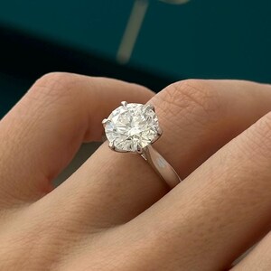 May include: A close-up of a silver engagement ring with a large, round, clear diamond. The ring is on a person's finger.