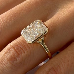 Op de afbeelding: Een gouden ring met een grote, rechthoekige, radiant-geslepen heldere edelsteen in een gladde zetting. De ringband is dun en van gepolijst goud, wat zorgt voor een strak en modern ontwerp dat de schittering van de edelsteen benadrukt.