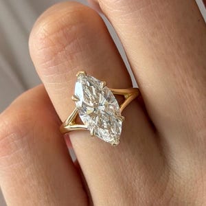 Op de afbeelding: Een gouden ring met een grote, transparante edelsteen in marquise-slijpvorm in een klauwzetting. De ringband heeft een gespleten ontwerp dat taps toeloopt naar de middelste steen, wat zorgt voor een strakke uitstraling van dit sieraad.