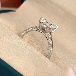 May include: A silver-toned ring featuring a large, round, faceted center stone held by a four-prong setting. The band is encrusted with small, clear stones along the shank. The ring is presented inside a dark teal box with the text NEXT DIAMOND.